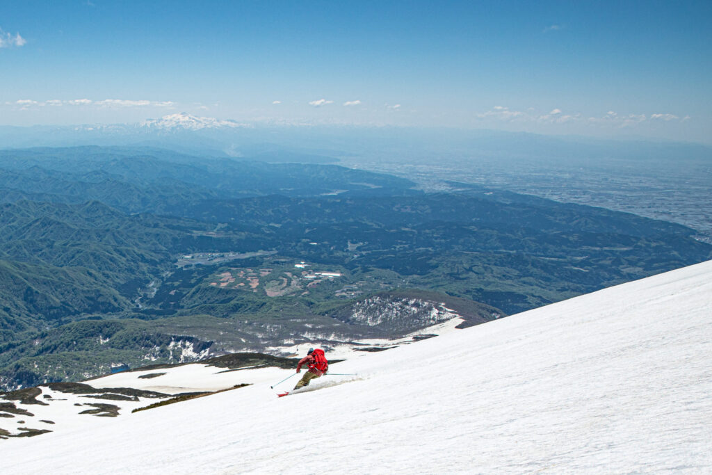 庄内平野と日本海を見下ろしながら、残雪期の鳥海山を滑るスキーヤー