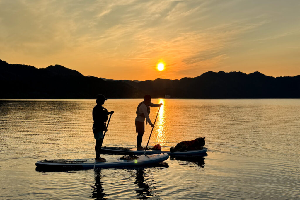 夕日に染まる田沢湖で、SUPを漕ぐ2人と秋田犬のシルエット