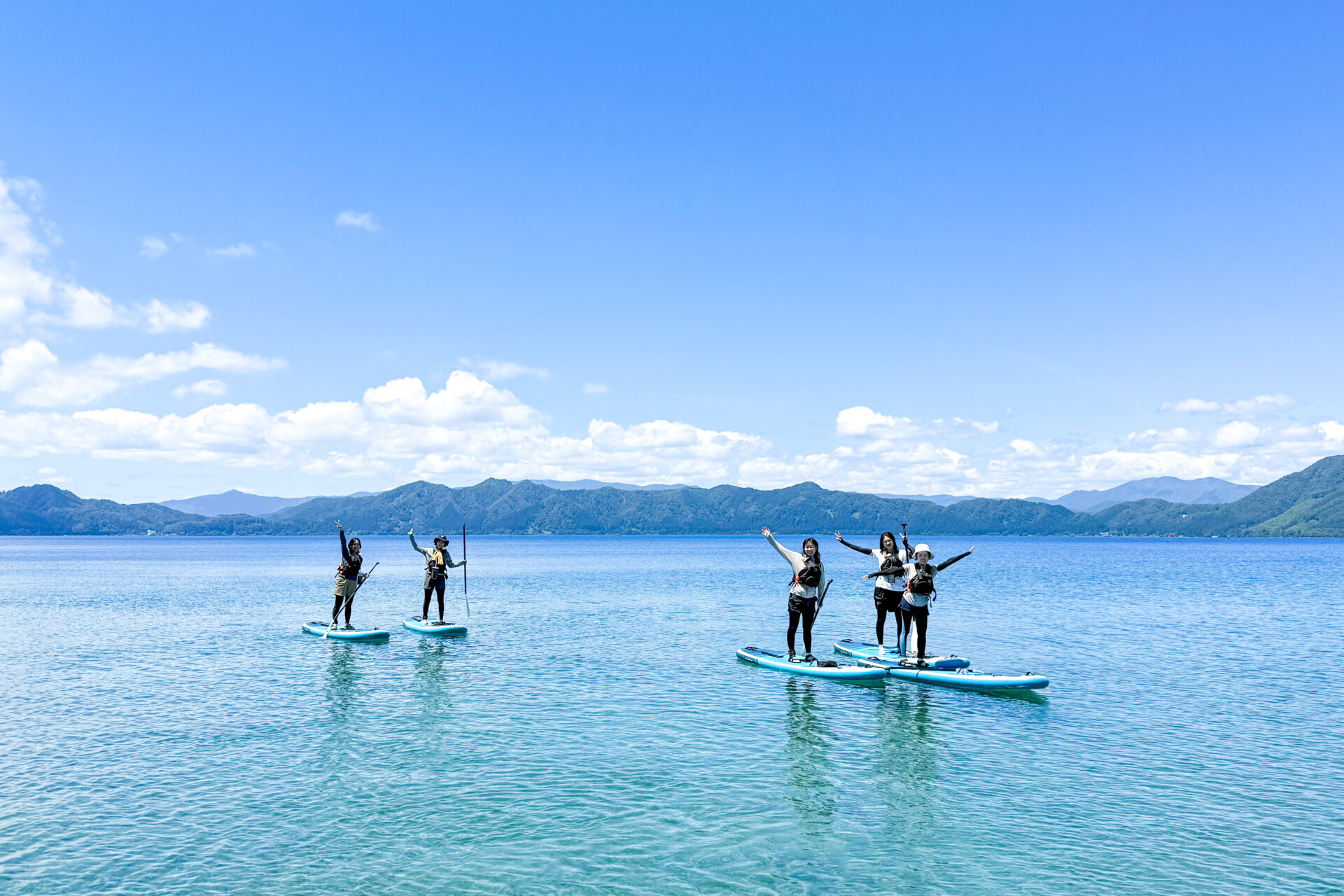 田沢湖の湖上でSUPに立ち手を上げる参加者と背景の山並み