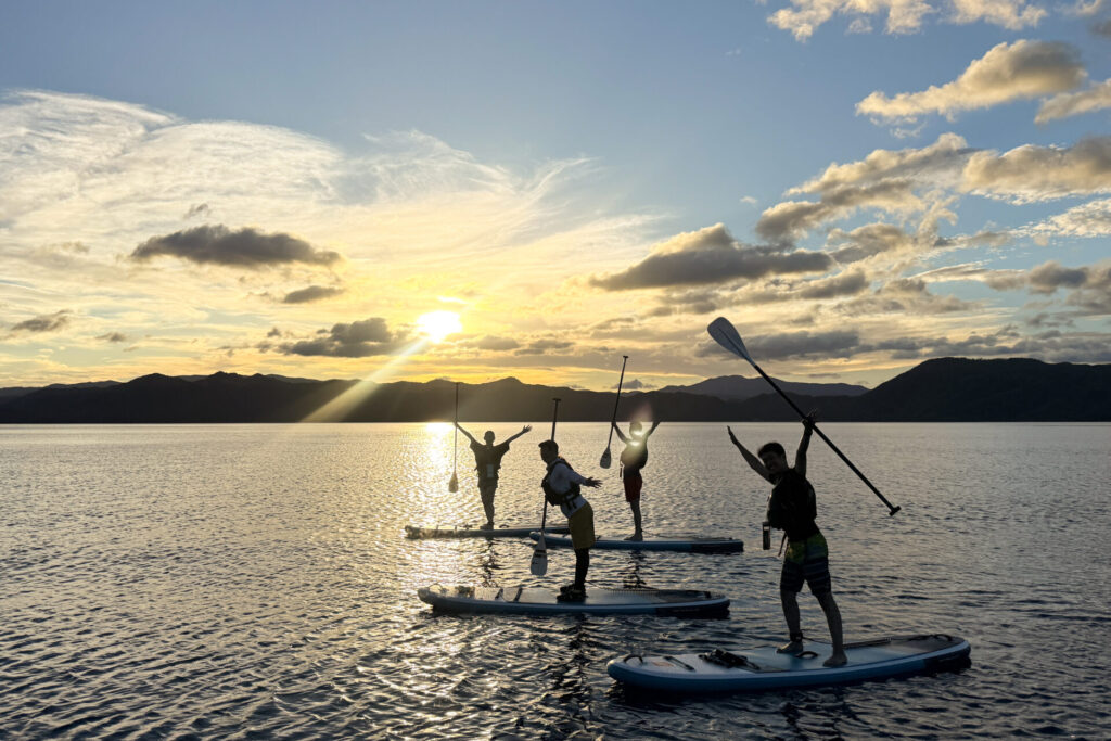沈む夕日と光の帯を背に田沢湖のSUPツアーでポーズを取る参加者たち
