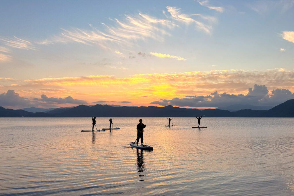 夕暮れ時の静かな田沢湖で、夕日に照らされたシルエットが美しいSUPクルージングの風景